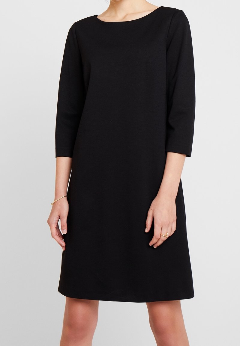 Femme portant une robe noire simple longueur genoux avec des manches trois-quarts, debout devant un fond gris clair uni.