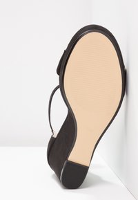 Chaussure à talon haut noire positionnée avec la semelle beige tournée vers le haut, talon et bride de cheville visibles, sur fond et mur blancs.