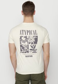 Kremkrāsas T-krekls ar grafisko dizainu uz aizmugures, kurā ir melni ziedu un putnu zīmējumi, kā arī teksts "ATYPICAL BACKYARD."