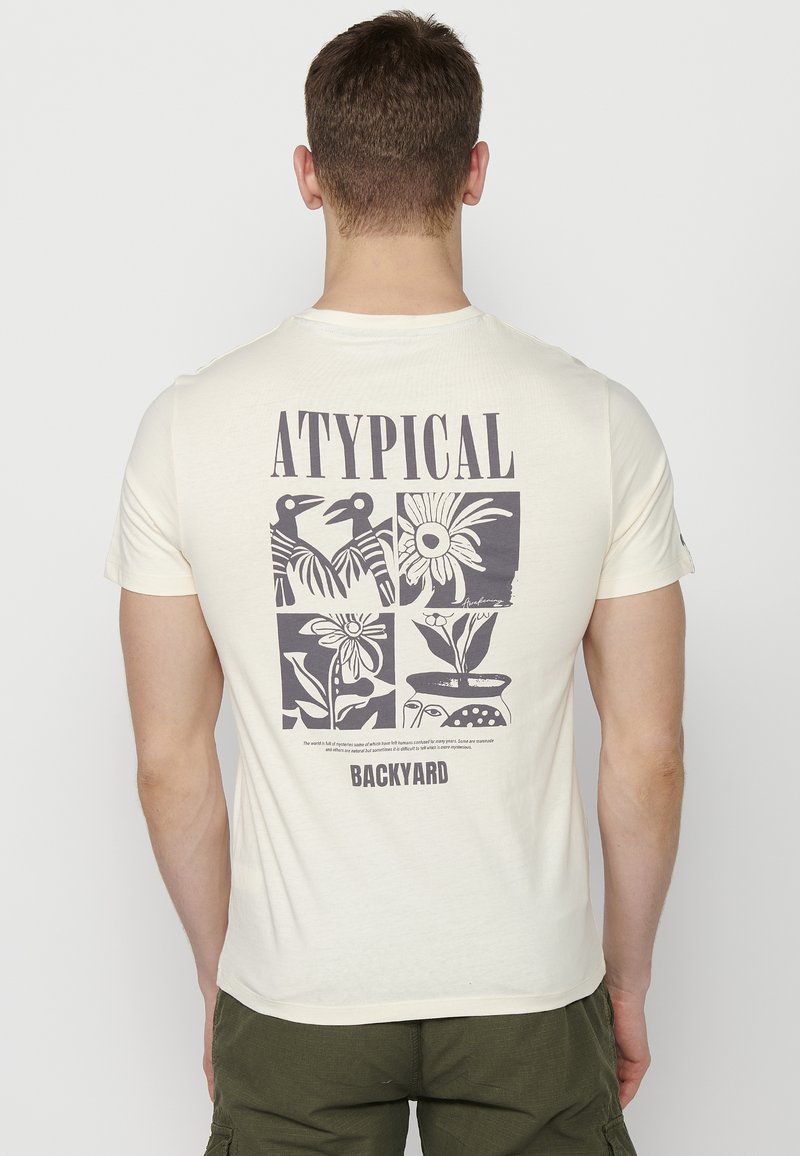 Kremkrāsas T-krekls ar grafisko dizainu uz aizmugures, kurā ir melni ziedu un putnu zīmējumi, kā arī teksts "ATYPICAL BACKYARD."