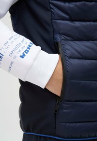 Marineblauer Puffer-Weste mit ge quilteter Textur, ausgestattet mit einer schwarzen Reißverschlusstasche. Der weiße Sweatshirtbund hat blauen Drucktext und gerippte Textur.