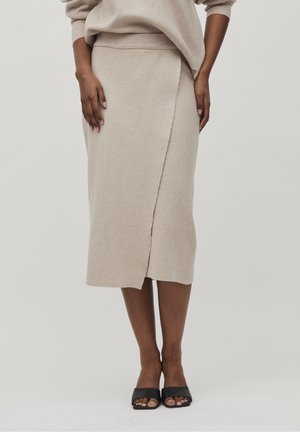Kvinde iført en beige omslagsmidi-nederdel kombineret med en matchende top og sorte åbne hælede sandaler står mod en ensfarvet baggrund.
