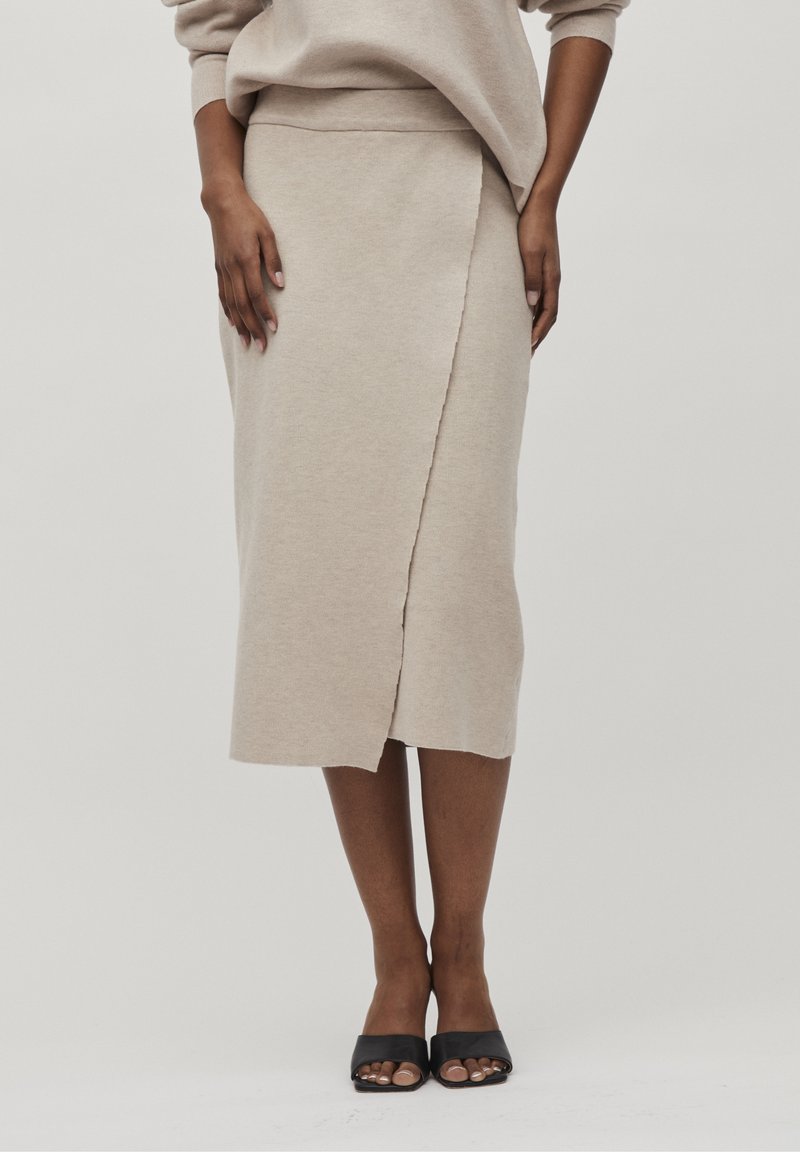 Femme portant une jupe midi beige portefeuille associée à un haut assorti et des sandales à talons noires à bout ouvert, debout devant un fond uni.