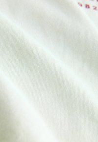 Tissu en coton blanc avec une texture côtelée, présentant des plis délicats et un petit détail imprimé en bordeaux. Surface douce et lisse.
