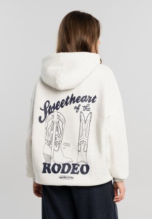 Henkilö, joka pukeutunut vaaleanväriseen huppariin, jossa on cowboy-saapasaiheisia kuvia ja teksti "Sweetheart of the Rodeo" takana, selin kameraan.