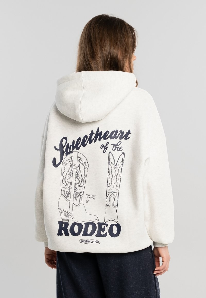 Personne portant un sweat à capuche de couleur claire avec des illustrations de bottes de cowboy et le texte « Sweetheart of the Rodeo » au dos, dos à la caméra.