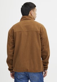 Brun fleecejakke med høy krage, avslappet passform og synlige sømdetaljer. Båret over blå jeans, som viser en teksturert overflate.