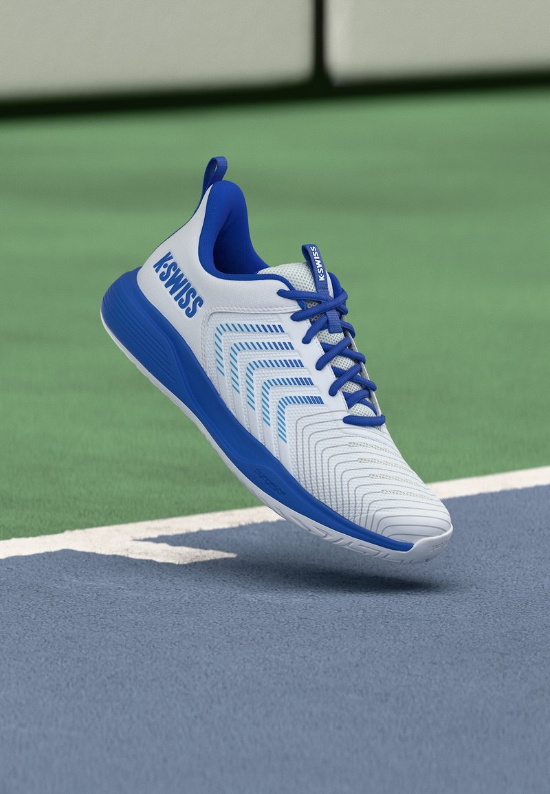 Λευκό και μπλε αθλητικό παπούτσι K-Swiss που αιωρείται πάνω από ένα μπλε και πράσινο γήπεδο τένις κοντά στη λευκή γραμμή ορίου.