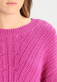 Stickad rosa tröja med ribbad rundhals och texturerade mönster, som visar upp varierande stygn och högkvalitativt garn.