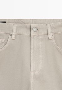 Pantalon en denim beige avec fermeture à boutons sur le devant, passants de ceinture et deux poches avant avec rivets métalliques.