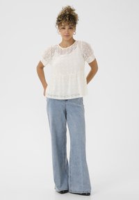 Haut en dentelle blanche avec de courtes manches bouffantes et une taille peplum, accompagné de jeans en denim évasés bleu clair. Design simple, tissu texturé.