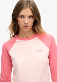 Jonge vrouw met lichtbruin haar die een roze en lichtroze SuperDry lange mouwen shirt draagt, recht vooruit kijkt met een neutrale uitdrukking.
