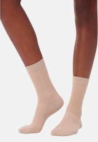 Dunkelhäutige Beine tragen hellbeige Crew-Socken mit subtilen Zickzack-Strickmustern und stehen vor einem schlichten weißen Hintergrund.
