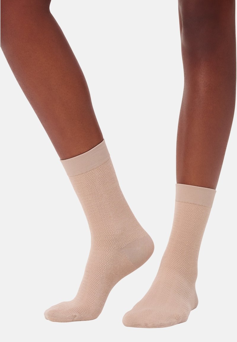Dunkelhäutige Beine tragen hellbeige Crew-Socken mit subtilen Zickzack-Strickmustern und stehen vor einem schlichten weißen Hintergrund.
