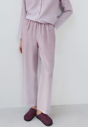 Person trägt lockere, staubig-rosa Hose und passendes Hemd mit Knöpfen, steht mit einer Hand hinter dem Rücken und trägt violette Slip-on-Schuhe.