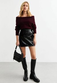 Suéter burdeos de hombros descubiertos, falda corta de cuero negra, botas altas negras y un pequeño bolso negro. Textura suave y diseño ajustado.