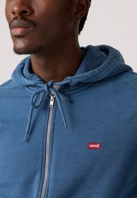 Blauwe zip-up hoodie van zacht materiaal, met een trekkoord in de capuchon en een rode Levi's logopatch op de borst. Zilveren ritsdetails.