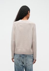 Mujer con pelo rizado largo, vestida con un suéter de punto beige y jeans azules descoloridos, vista desde atrás sobre un fondo liso.