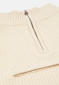 Maglione a costine di colore crema con colletto alto e zip corta. Presenta una texture morbida e un delicato dettaglio di cucitura lungo la zip.