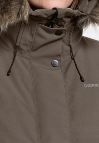 Braune Winterjacke mit Fellkapuze, schwarzen Kordeln und Druckknopfverschluss am Hals, die einen Teil des Didriksons-Logos zeigt.