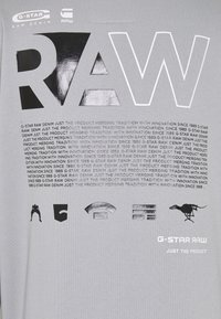 Camiseta gris con grandes letras negras "RAW", varios gráficos pequeños y texto repetido sobre la tradición e innovación de G-Star Raw.