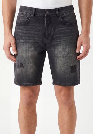 Hombre con pantalones cortos de denim negros desteñidos con parches desgastados, de pie frente a un fondo blanco liso.