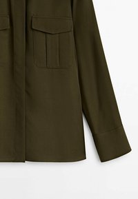 Olive green shirt featuring a pointed collar, two chest pockets, long sleeves, and a straight hem. Made from a smooth, lightweight fabric.