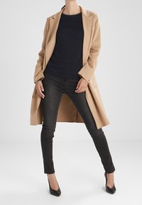 Beige ullkappa med ficklappar, marinblå tröja, svarta smala jeans och svarta spetsiga klackar. Slät textur och skräddarsydd passform.