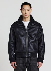 Uomo che indossa una giacca bomber nera con zip e colletto in pelliccia, una camicia bianca sotto e jeans neri, in piedi davanti a uno sfondo semplice.