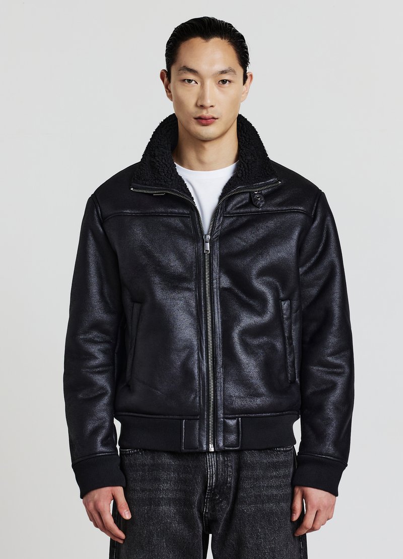 Uomo che indossa una giacca bomber nera con zip e colletto in pelliccia, una camicia bianca sotto e jeans neri, in piedi davanti a uno sfondo semplice.