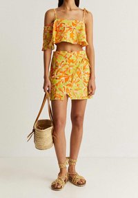 Top court floral jaune avec un ourlet froncé, associé à une jupe portefeuille assortie. Les accessoires comprennent un sac en paille tressée et des sandales tressées.