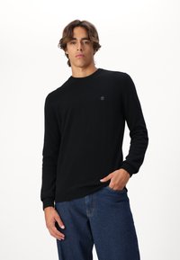 Schwarzer Strickpullover mit Rundhalsausschnitt, gerippten Bündchen und kleinem Logo auf der Brust, kombiniert mit blauen Jeans. Weiche Textur und figurbetonter Schnitt.