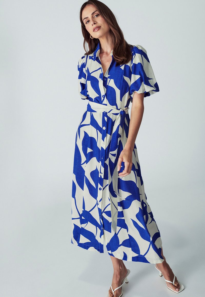 Šviesiai ruda midi suknelė su mėlynu lapų raštu, trumpomis pūstomis rankovėmis ir surišamu juosmeniu. Medžiaga atrodo lengva ir plazdanti.