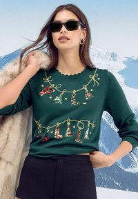 Zelena pulover s valovitim izrezom, okrašen s sijočimi črkami in drevesci v prazničnem slogu, v kombinaciji z črno krilom in krznenim plaščem.