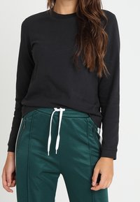 Maglietta nera a maniche lunghe abbinata a pantaloni verdi con vita elastica, caratterizzati da lacci bianchi e tasche laterali con zip.