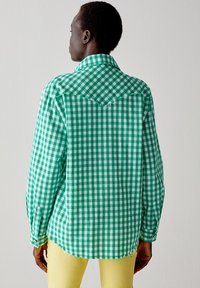 Camicia a scacchi verde e bianca con colletto, maniche lunghe e orlo sfilacciato. Il tessuto è leggero e dalla consistenza morbida.