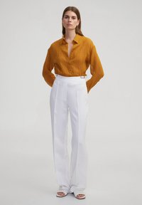 Senfgelbe, zerknitterte Bluse mit Kragen und Knopfverschluss, kombiniert mit hochtaillierten weißen Hosen mit silbernen Hardware-Akzenten.