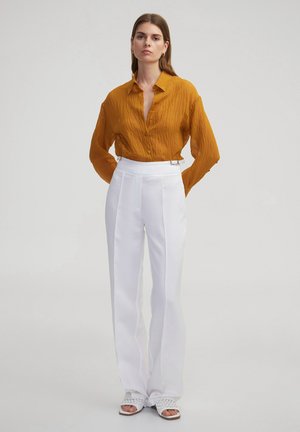 Blusa arrugada de color mostaza con cuello y cierre de botones, combinada con pantalones blancos de talle alto con acentos de hardware plateado.