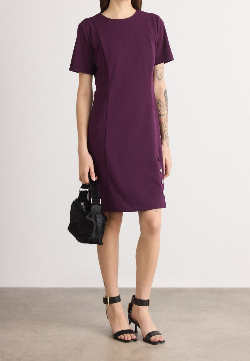 Femme portant une robe violette longueur genou avec des boutons sur les côtés, des talons noirs à bride à la cheville, tenant un petit sac à main noir.