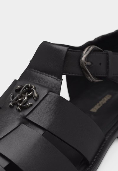 Sandale en cuir noir avec larges brides, fermeture à boucle métallique, et un logo décoratif en métal sur la bride supérieure.