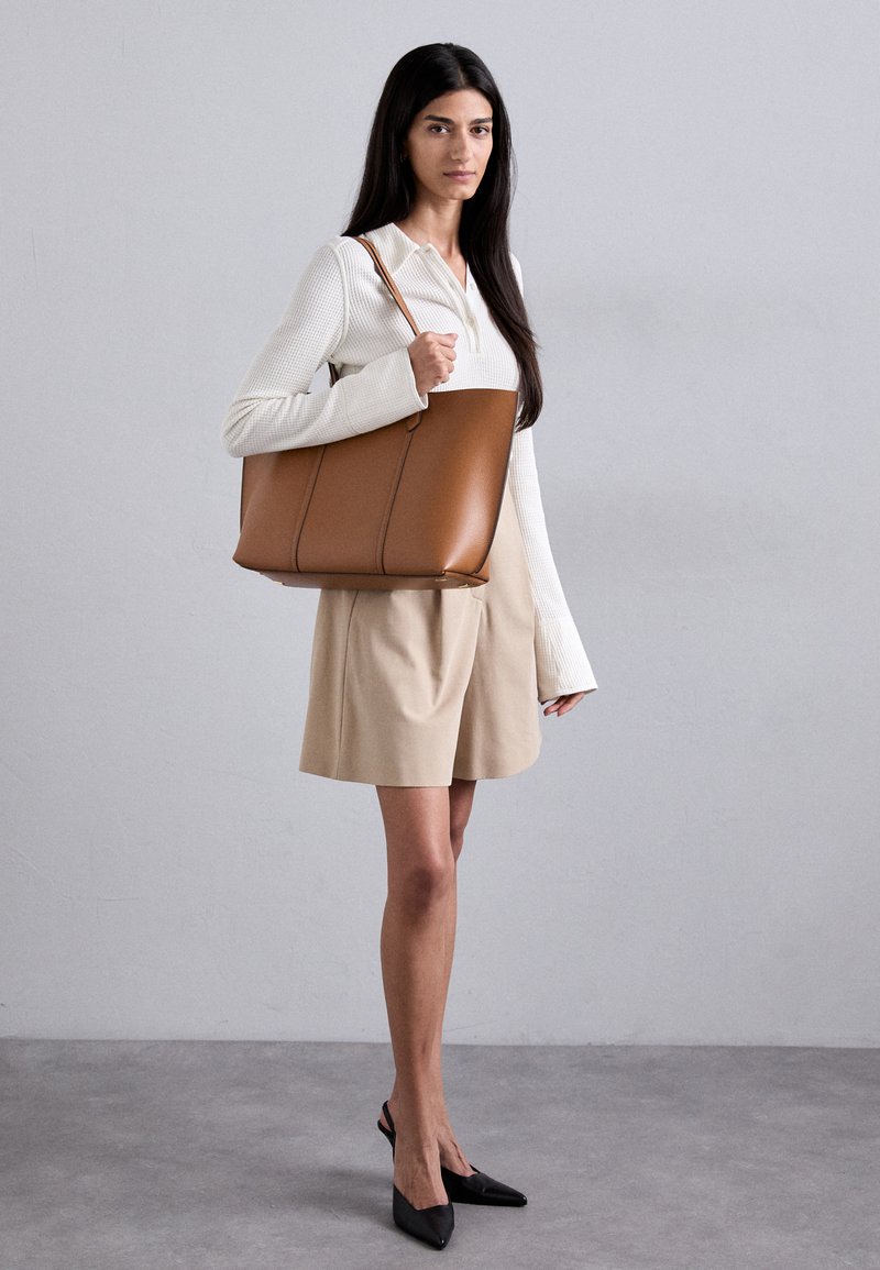 Sac fourre-tout en cuir marron au design structuré, doté de deux poignées supérieures et de coutures visibles, associé à un haut à manches longues blanc et à un short beige.