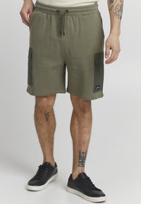 Shorts en coton vert olive avec une taille élastique, un cordon de serrage et des poches cargo latérales. Patch logo noir sur la jambe inférieure.