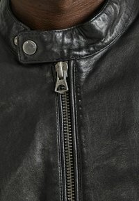 Giacca da moto in pelle nera con colletto alto, chiusura con zip argentata e dettagli con bottoni a pressione. Texture liscia con leggera lucentezza.
