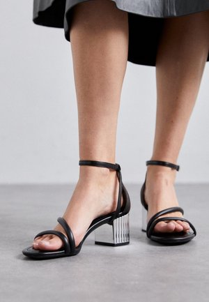 Femme portant des sandales noires à talons hauts avec brides et talons blocs métalliques, debout sur un sol gris, ourlet de jupe visible.