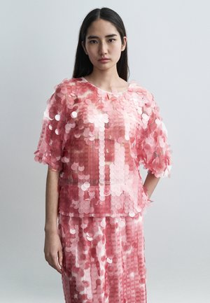 Różowa cekinowa bluzka z krótkimi rękawami, nałożona na dopasowaną spódnicę. Posiada okrągły dekolt oraz przezroczystą tkaninę bazową o połyskującej fakturze.