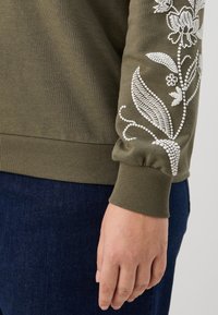 Felpa verde oliva con ricamo floreale bianco sulla manica, caratterizzata da polsini e fondo a coste, realizzata in morbido tessuto misto cotone.