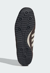 Potplat crnog Adidas tenisica s kružnim šiljcima za prianjanje, horizontalnim utorima i bež dizajnom s tri pruge sa strane.
