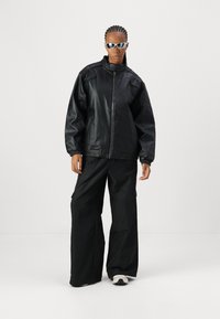 Veste noire en synthétique avec col montant et poignets élastiques, associée à un pantalon cargo noir ample. Complétée par des lunettes de soleil réfléchissantes.