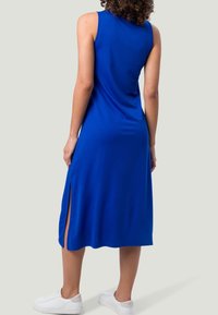 Sininen hihaton mekko, joka on valmistettu sileästä kankaasta, sivuhalkioilla ja keskipituisena, yhdistettynä valkoisiin tenniskenkiin.