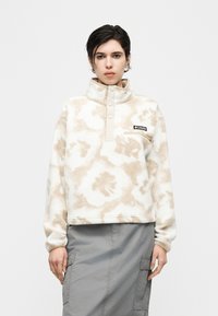 Άτομο που φοράει μπεζ και λευκό φλις πουλόβερ με κουμπιά και γκρι φούστα τύπου cargo, στέκεται μπροστά σε απλό φόντο.